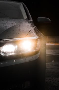 Glistening raindrops on a car headlight illuminate the night scene vividly.